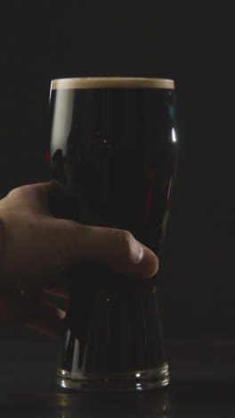 Vertical-Video-Of-Person-Picking-Up-Pint-Of-Irish-Stout-In-Glass-Against-Black-Studio-Background-To-Celebrate-St-Patricks-Day